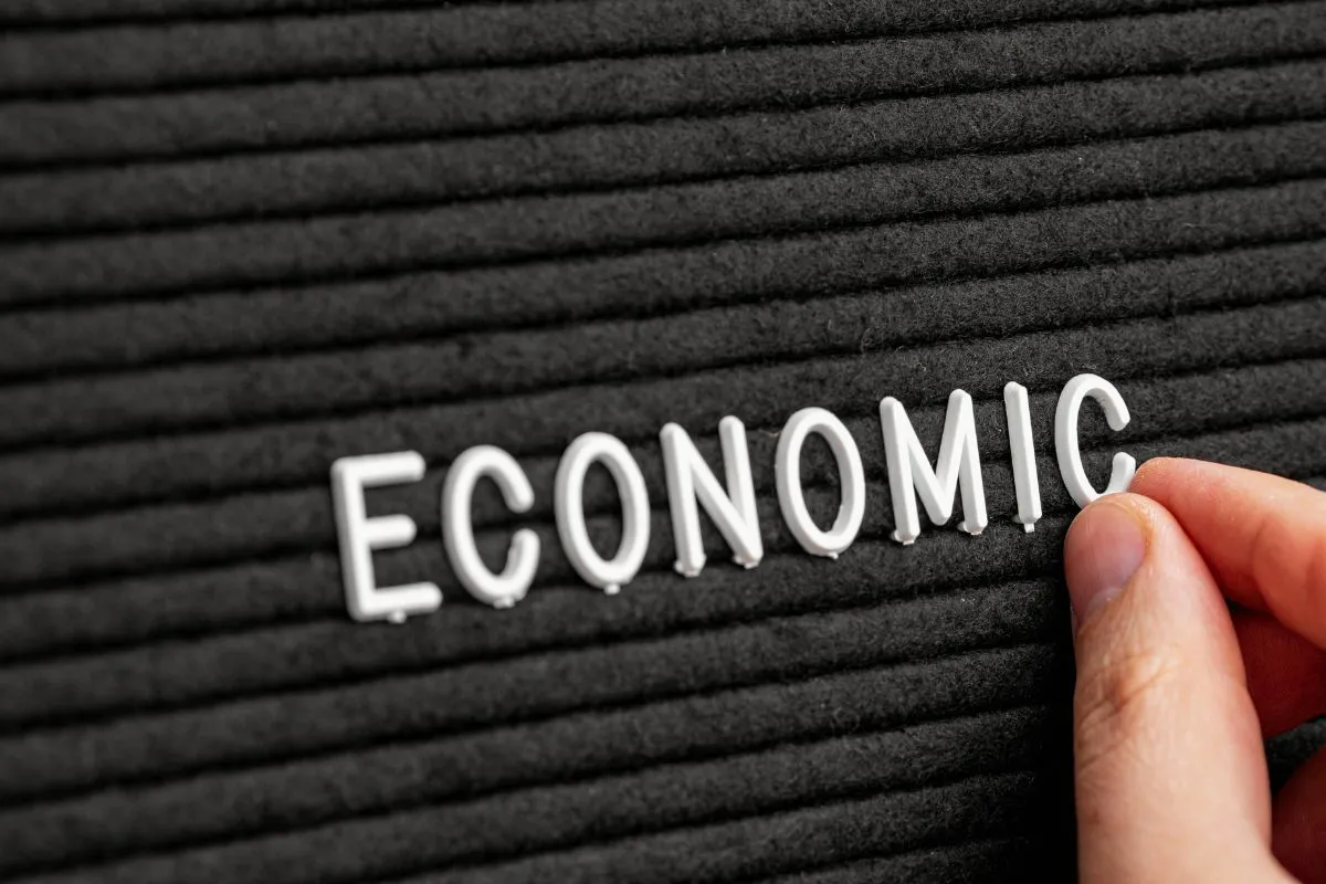 Mão posicionando letra na palavra Economic em painel preto, representando economia, mercado financeiro e análise econômica.