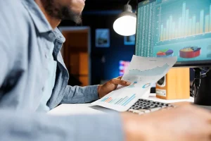 Homem analisando gráficos financeiros no computador enquanto segura relatório com dados econômicos