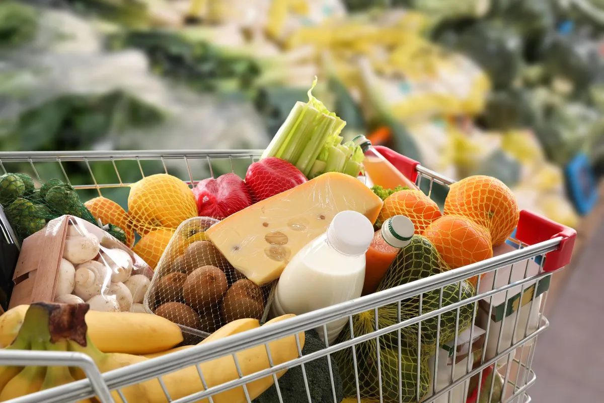 Carrinho de supermercado cheio com alimentos como frutas, leite e queijo representando cesta básica e custo de vida