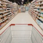 Carrinho de supermercado vazio em corredor com prateleiras cheias representando aumento do custo da cesta básica