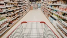 Carrinho de supermercado vazio em corredor com prateleiras cheias representando aumento do custo da cesta básica