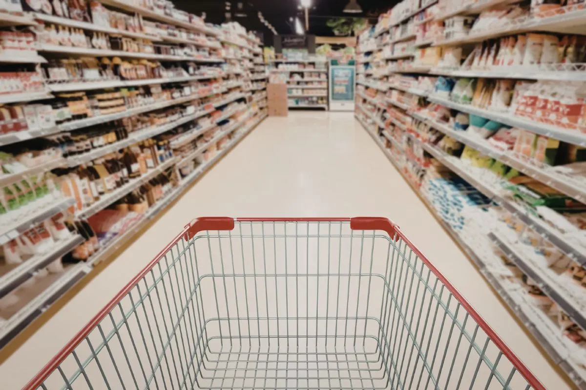 Carrinho de supermercado vazio em corredor com prateleiras cheias representando aumento do custo da cesta básica