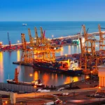 Porto com guindastes, navios cargueiros e contêineres representando comércio internacional e transporte marítimo
