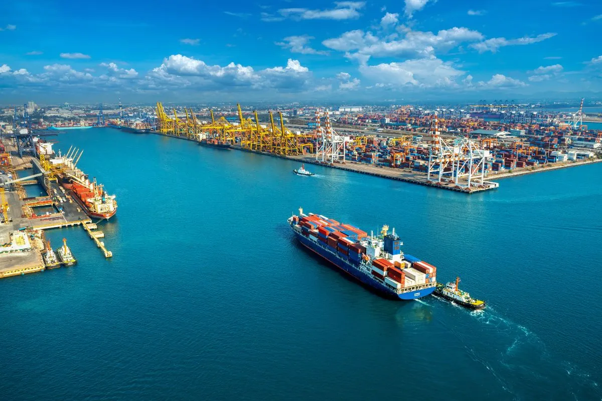 Navio de contêineres navegando em porto industrial com guindastes e terminal de cargas ao fundo