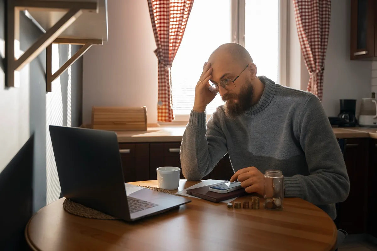 Homem preocupado usando calculadora com moedas e laptop na mesa representando dificuldades financeiras