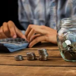 Pessoa usando calculadora ao lado de moedas empilhadas e pote com dinheiro representando economia e planejamento financeiro