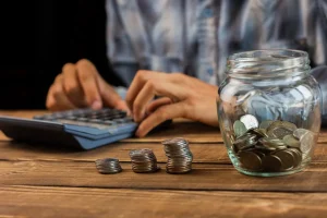 Pessoa usando calculadora ao lado de moedas empilhadas e pote com dinheiro representando economia e planejamento financeiro