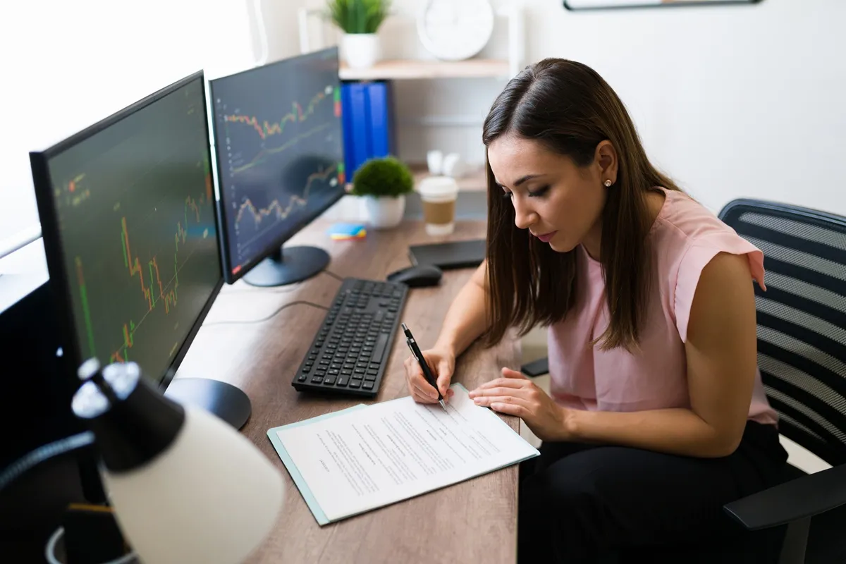 Mulher analisando gráficos financeiros em dois monitores enquanto assina documento no escritório em casa