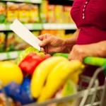 Pessoa segurando lista de compras ao lado de carrinho com frutas e alimentos no supermercado