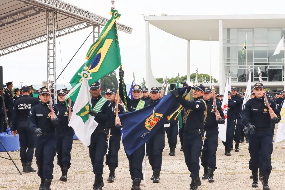 Policiais militares marchando em formação durante desfile cívico com bandeira do Brasil e estandartes oficiais