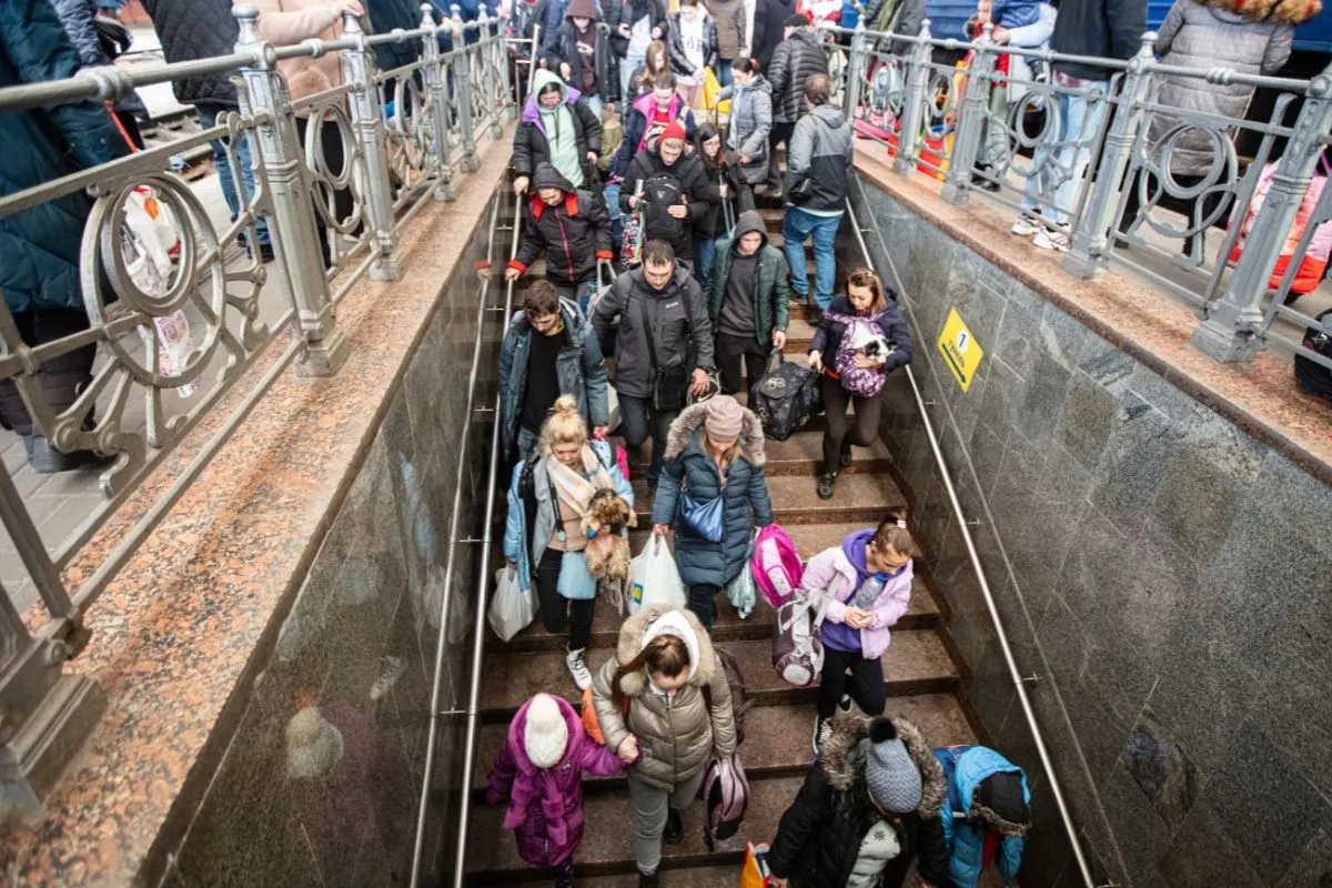 Pessoas descendo escadas de metrô com malas e bolsas em contexto de migração ou refúgio