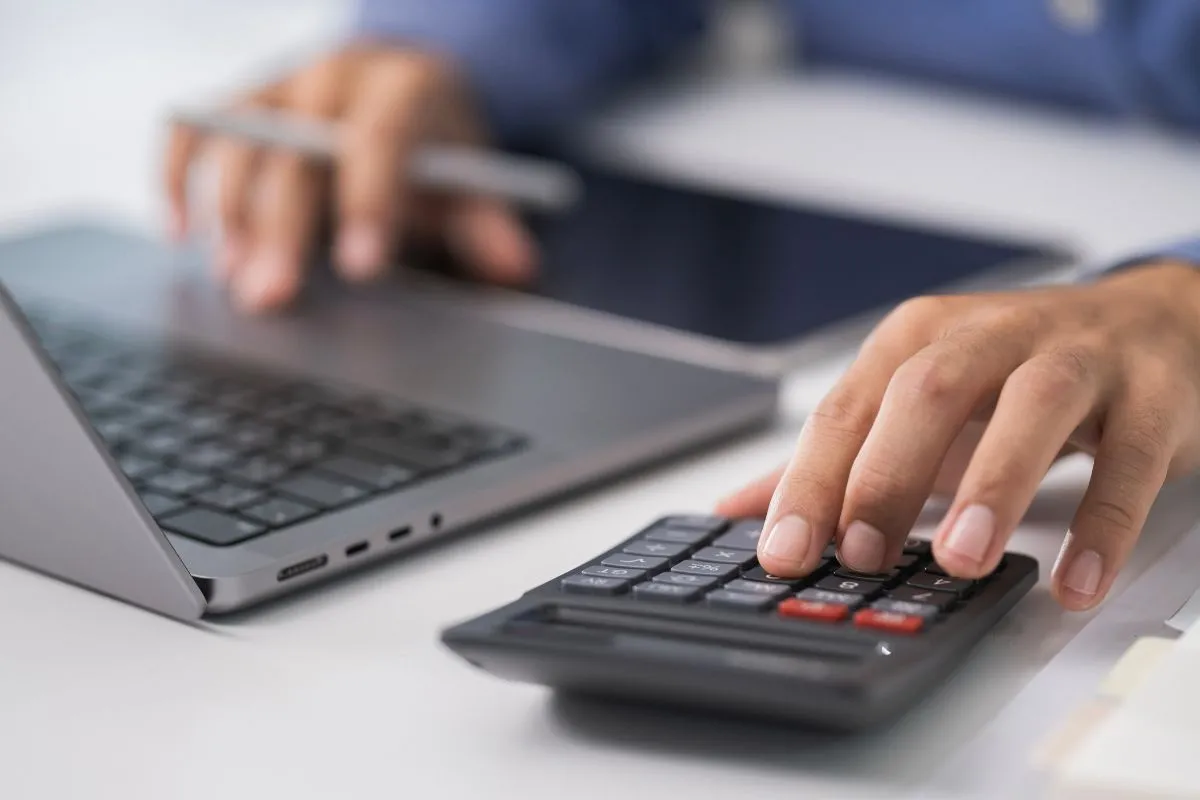 Pessoa utilizando calculadora ao lado de notebook durante cálculo de impostos e despesas financeiras
