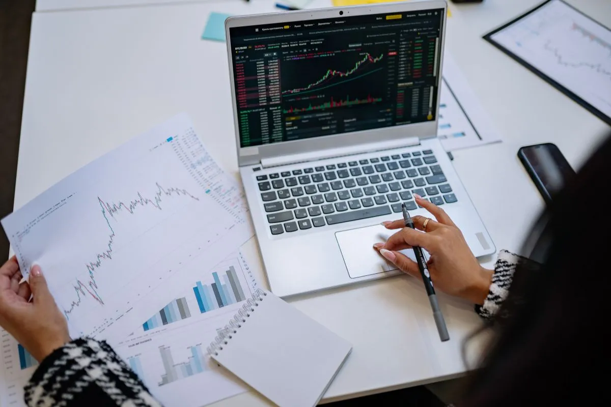 Pessoa analisando gráficos financeiros no notebook e em planilhas para comparar indicadores econômicos.