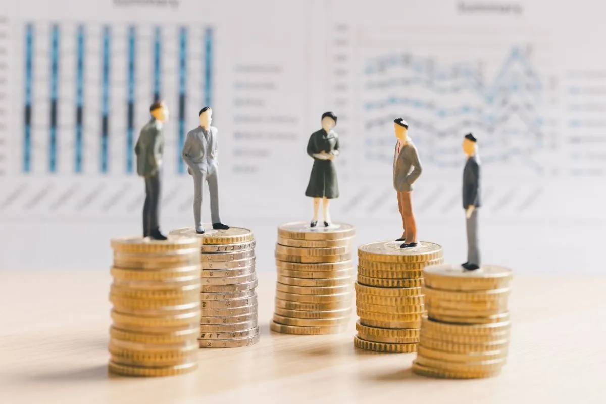 Miniaturas de pessoas sobre pilhas de moedas representando arrecadação, distribuição de renda e gastos públicos