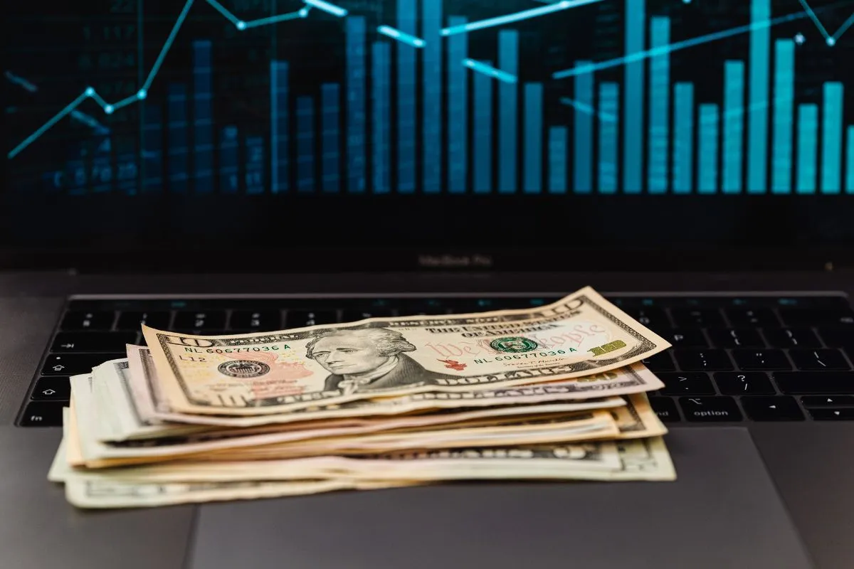 Notas de dólar sobre notebook com gráfico financeiro ao fundo representando investimentos