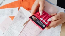 Pessoa usando calculadora sobre recibos e contas representando impacto dos impostos no preço final