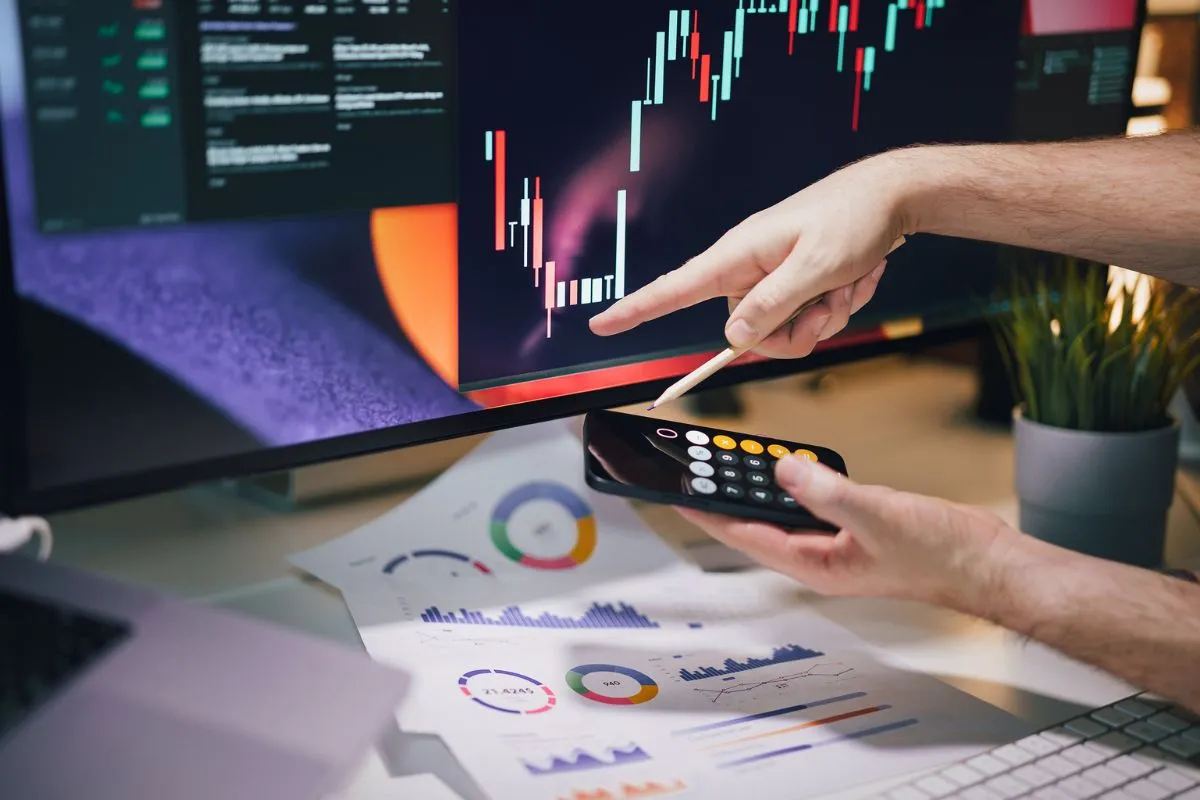Pessoa analisando gráfico de mercado financeiro em monitor enquanto usa calculadora para investimentos