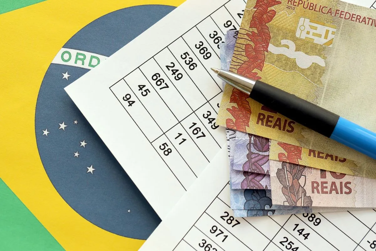 Notas de real sobre planilha financeira com caneta e bandeira do Brasil ao fundo