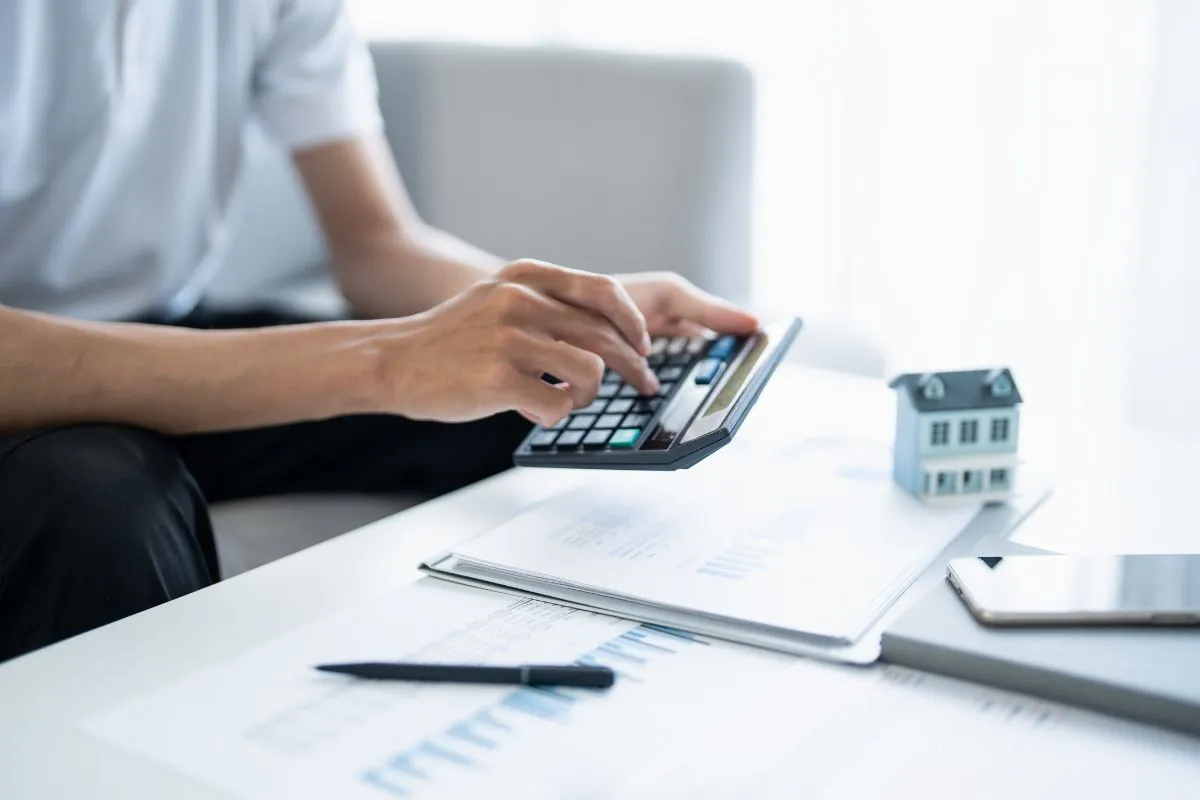 Pessoa usando calculadora com documentos financeiros e miniatura de casa sobre a mesa