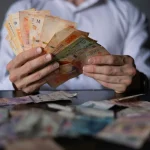 Pessoa segurando e contando notas de real brasileiro sobre mesa com dinheiro espalhado