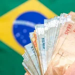 Mão segurando notas de real brasileiro com bandeira do Brasil desfocada ao fundo