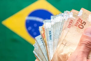 Mão segurando notas de real brasileiro com bandeira do Brasil desfocada ao fundo
