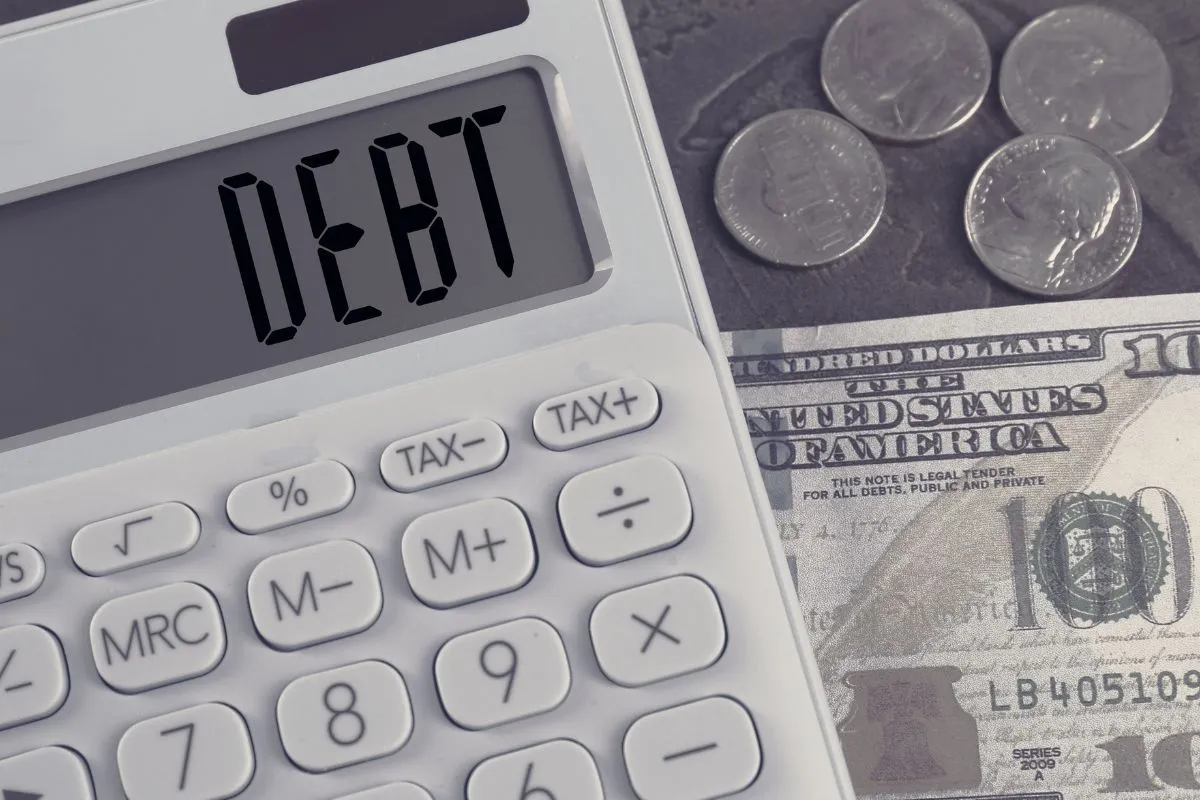 Calculadora exibindo a palavra debt ao lado de notas e moedas relacionada aos juros da dívida pública