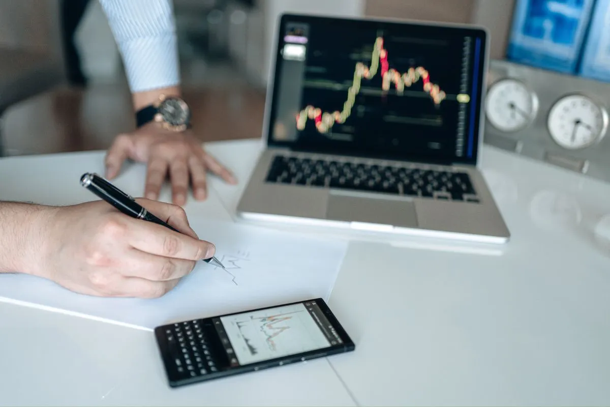 Profissional analisando gráficos financeiros em notebook e celular durante acompanhamento de mercado econômico.