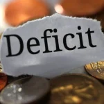 Palavra deficit sobre moedas representando rombo fiscal e desequilíbrio nas contas públicas brasileiras