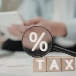 Lupa destacando símbolo de porcentagem sobre blocos com palavra tax em cenário de análise tributária.