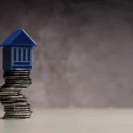Miniatura de prédio sobre pilha de moedas simbolizando teto de gastos e limite das despesas públicas.