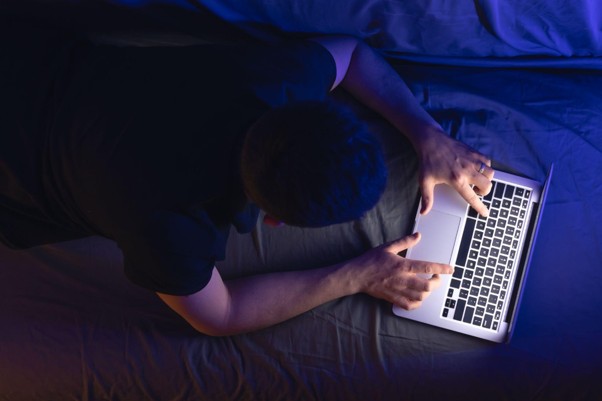 Visão aérea de um homem deitado na cama no escuro, usando um notebook que reflete luz azul em seu rosto.