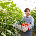 Produtora rural segurando caixa com legumes em estufa agrícola ligada ao abastecimento e preço dos alimentos.