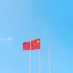 Fotografia em plano aberto e ângulo contra-plongée, capturando a bandeira da China hasteada em um mastro metálico central. A bandeira, de cor vermelha vibrante, exibe cinco estrelas amarelas no canto superior esquerdo. Ao lado, dois mastros adicionais estão vazios. O fundo é composto inteiramente por um céu azul-celeste claro, límpido e sem nuvens, realçando o contraste cromático entre o vermelho da bandeira e o azul do céu.