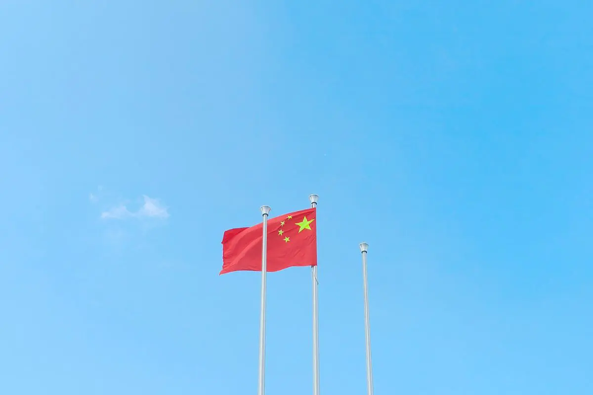 Fotografia em plano aberto e ângulo contra-plongée, capturando a bandeira da China hasteada em um mastro metálico central. A bandeira, de cor vermelha vibrante, exibe cinco estrelas amarelas no canto superior esquerdo. Ao lado, dois mastros adicionais estão vazios. O fundo é composto inteiramente por um céu azul-celeste claro, límpido e sem nuvens, realçando o contraste cromático entre o vermelho da bandeira e o azul do céu.