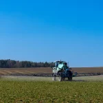 Vista traseira de um grande pulverizador agrícola azul e branco em movimento, com barras aplicadoras totalmente estendidas, liberando uma névoa de defensivos sobre uma lavoura verde sob céu azul limpo.