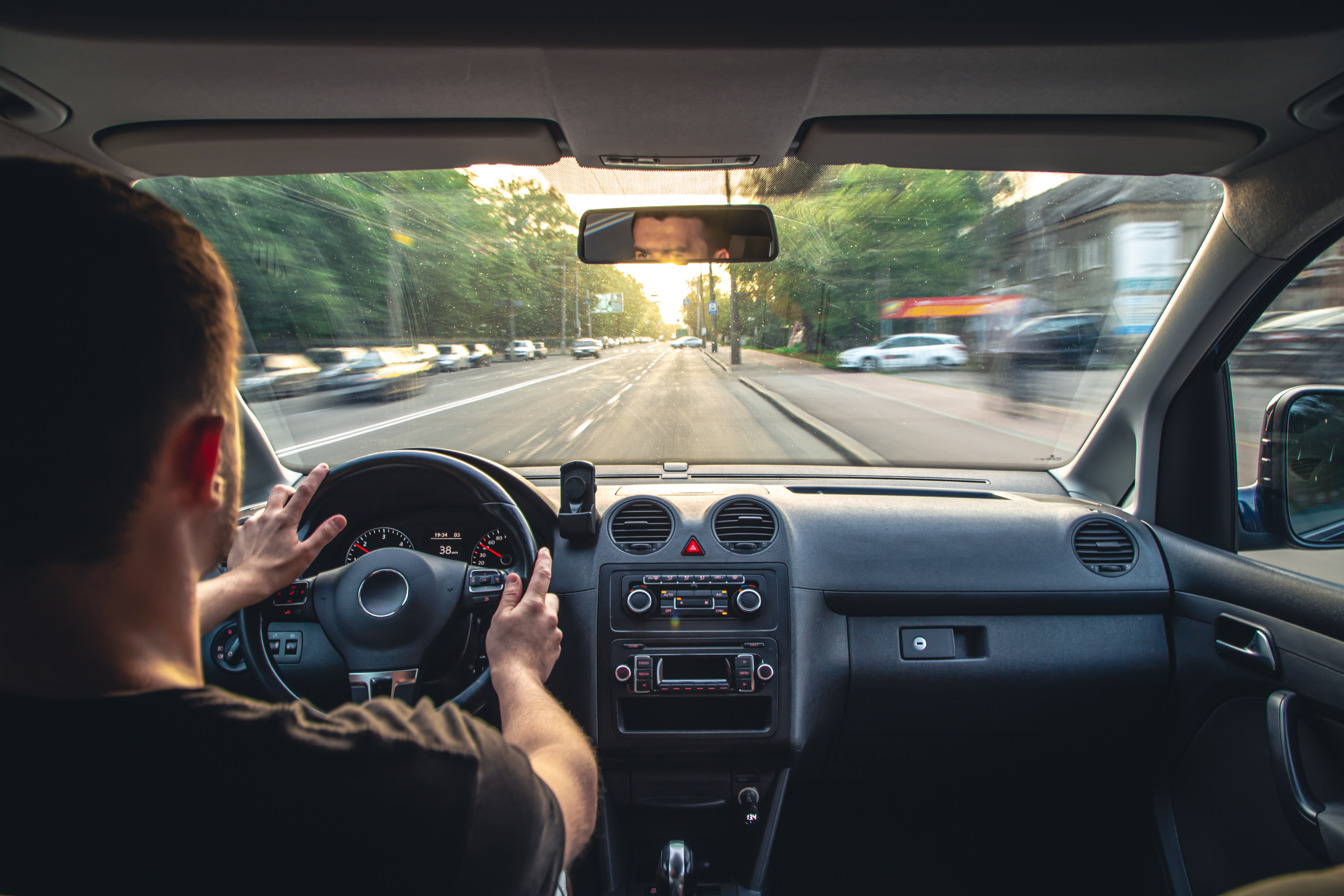 Visão de dentro do veículo mostrando um motorista de aplicativo com as duas mãos no volante, dirigindo por uma avenida da cidade durante o dia.