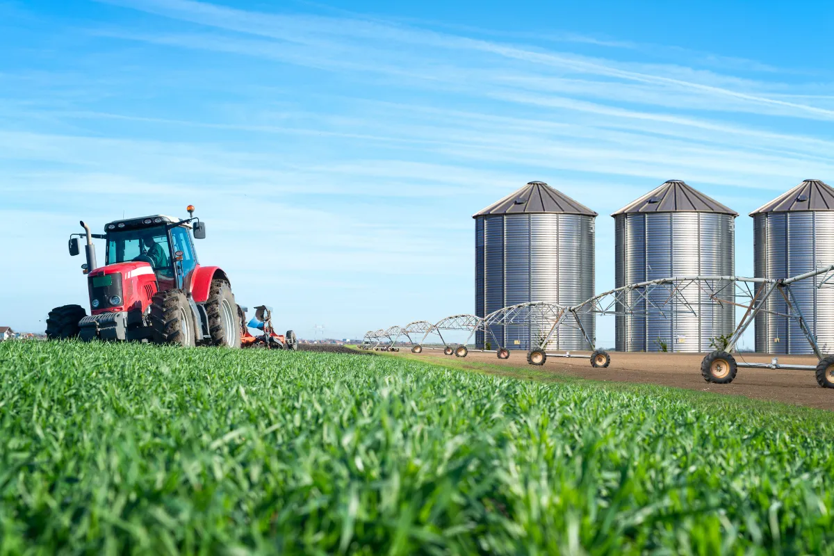 Trator trabalha próximo a silos metálicos e sistema de irrigação, estrutura essencial para armazenagem e logística agrícola.