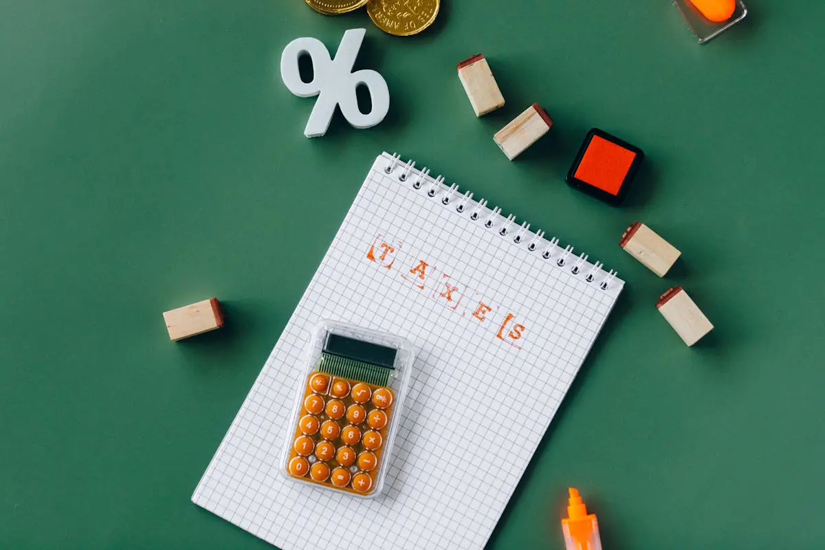 Vista superior em plano zenital de um caderno quadriculado com a palavra TAXES carimbada em vermelho, uma pequena calculadora com botões laranjas, um símbolo de porcentagem branco, moedas douradas e blocos de carimbo de madeira sobre uma mesa verde fosca.