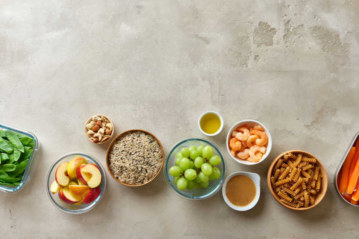 Ingredientes saudáveis como maçã, uvas, arroz integral, camarão, vegetais e massas dispostos sobre superfície clara.