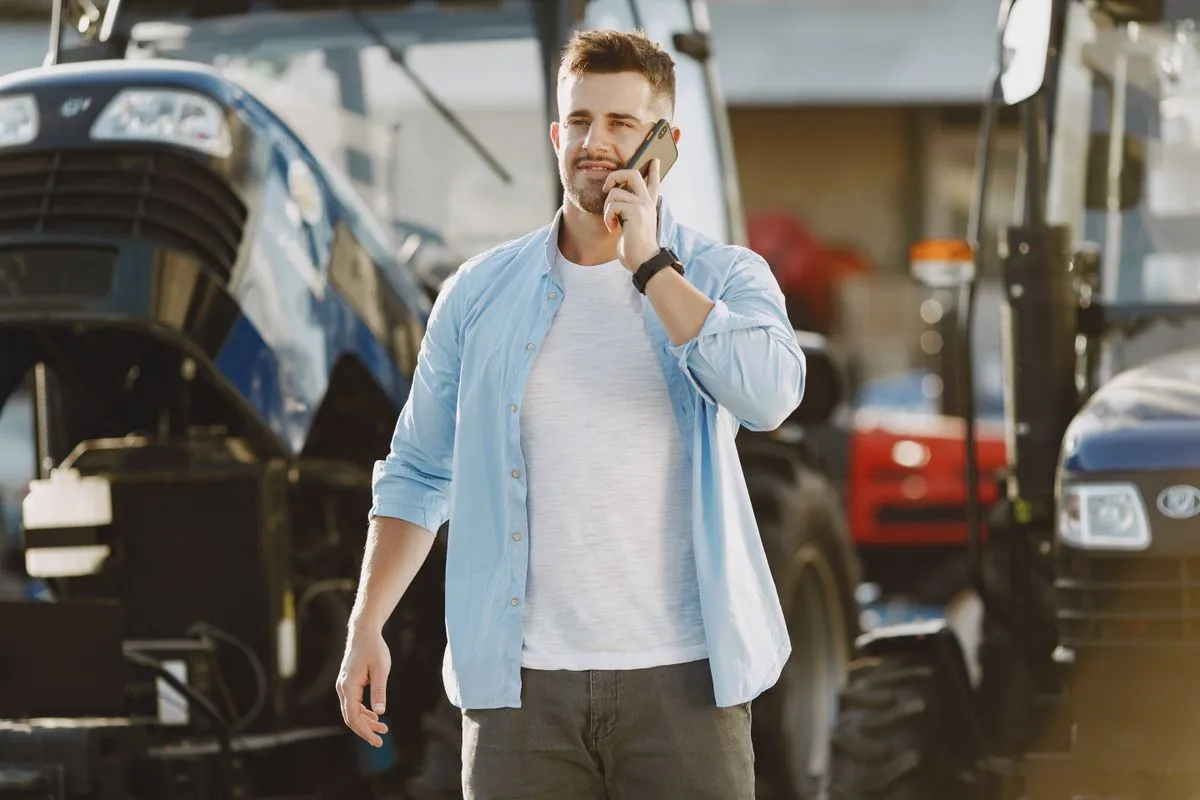 Homem jovem de pele clara e barba rala, vestindo camiseta branca e camisa de botão azul aberta, fala ao smartphone enquanto caminha em um pátio cercado por tratores agrícolas.