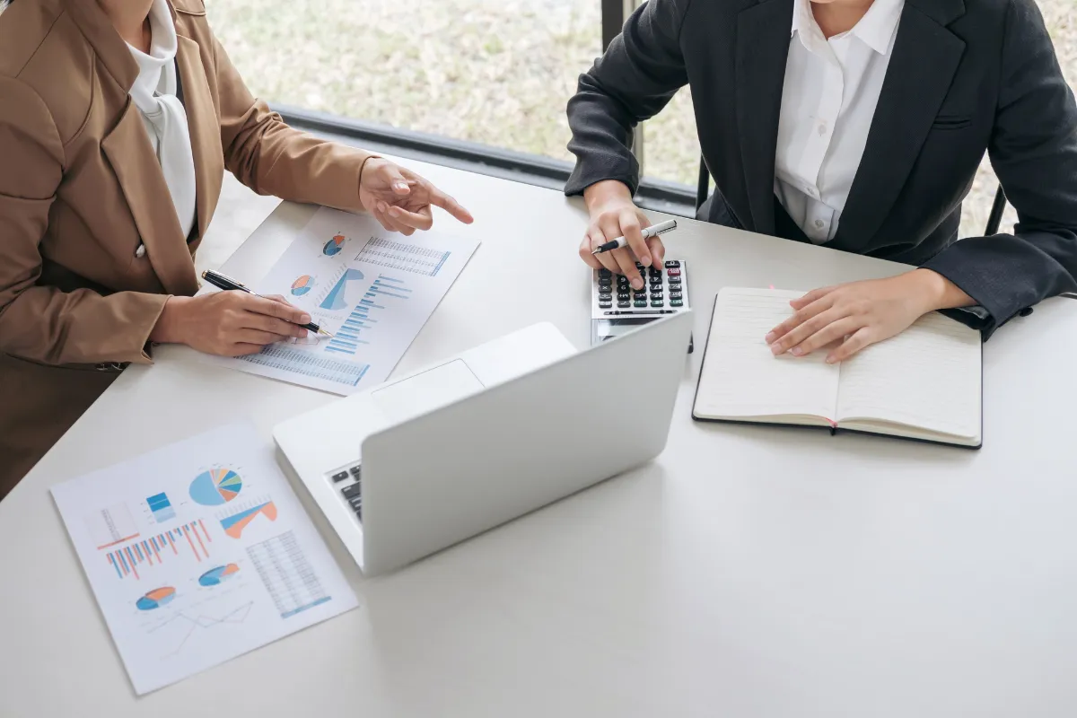 Duas profissionais analisando gráficos financeiros ao lado de um notebook e uma calculadora.