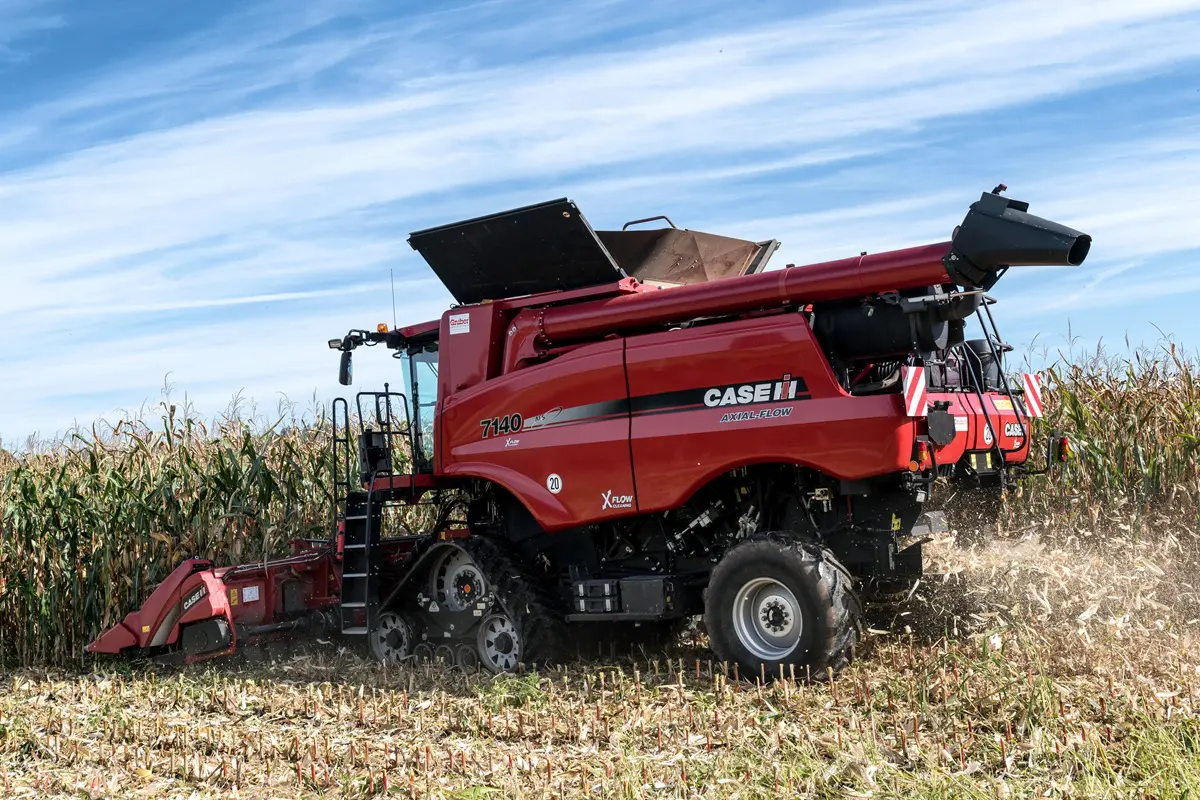 Fotografia em plano aberto e ângulo normal mostrando uma colheitadeira Case IH vermelha modelo 7140 Axial-Flow operando em uma plantação de milho. A máquina possui esteiras na parte dianteira e rodas na traseira, movendo-se sobre restos de cultura colhida. O céu azul com nuvens tênues aparece ao fundo, e a máquina está em plena atividade de colheita.