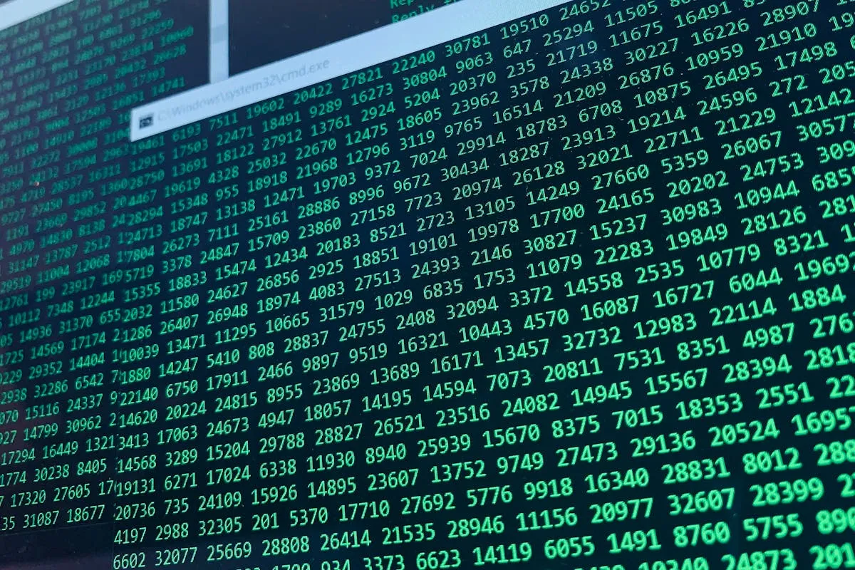 Close-up em ângulo diagonal de uma tela de computador exibindo centenas de sequências numéricas de cinco dígitos na cor verde brilhante sobre fundo preto, com uma janela sobreposta de prompt de comando do Windows.