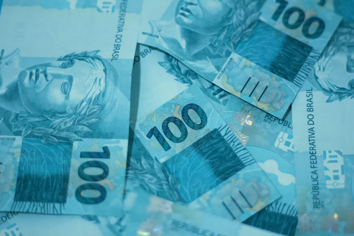 Close-up de várias cédulas brasileiras de cem reais espalhadas e sobrepostas sob uma iluminação fria e azulada, exibindo o número 100, a efígie da República e a inscrição República Federativa do Brasil.