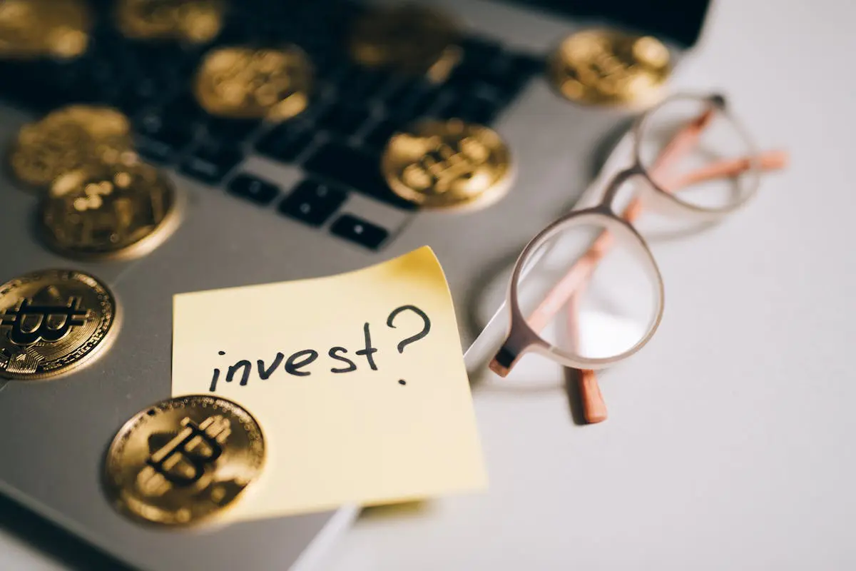 Vista superior aproximada em ângulo diagonal de um bloco de notas autoadesivo amarelo com a palavra invest escrita à mão ao lado de um ponto de interrogação, posicionado sobre um notebook cercado por moedas douradas com o símbolo do Bitcoin e óculos de armação fina.