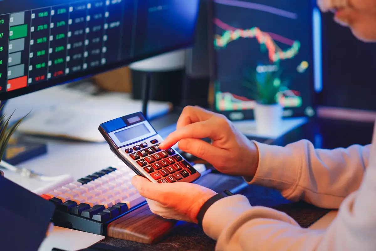 Fotografia em plano médio mostrando um operador financeiro trabalhando em um escritório tecnológico. Em primeiro plano, as mãos de uma pessoa operam uma calculadora de mesa, calculando margens de lucro. Ao fundo, dois monitores de computador exibem gráficos financeiros complexos com linhas de tendência em cores neon, verdes e vermelhas, indicando flutuações de mercado. O ambiente possui iluminação técnica e acessórios como um teclado gamer e uma pequena planta ornamental.