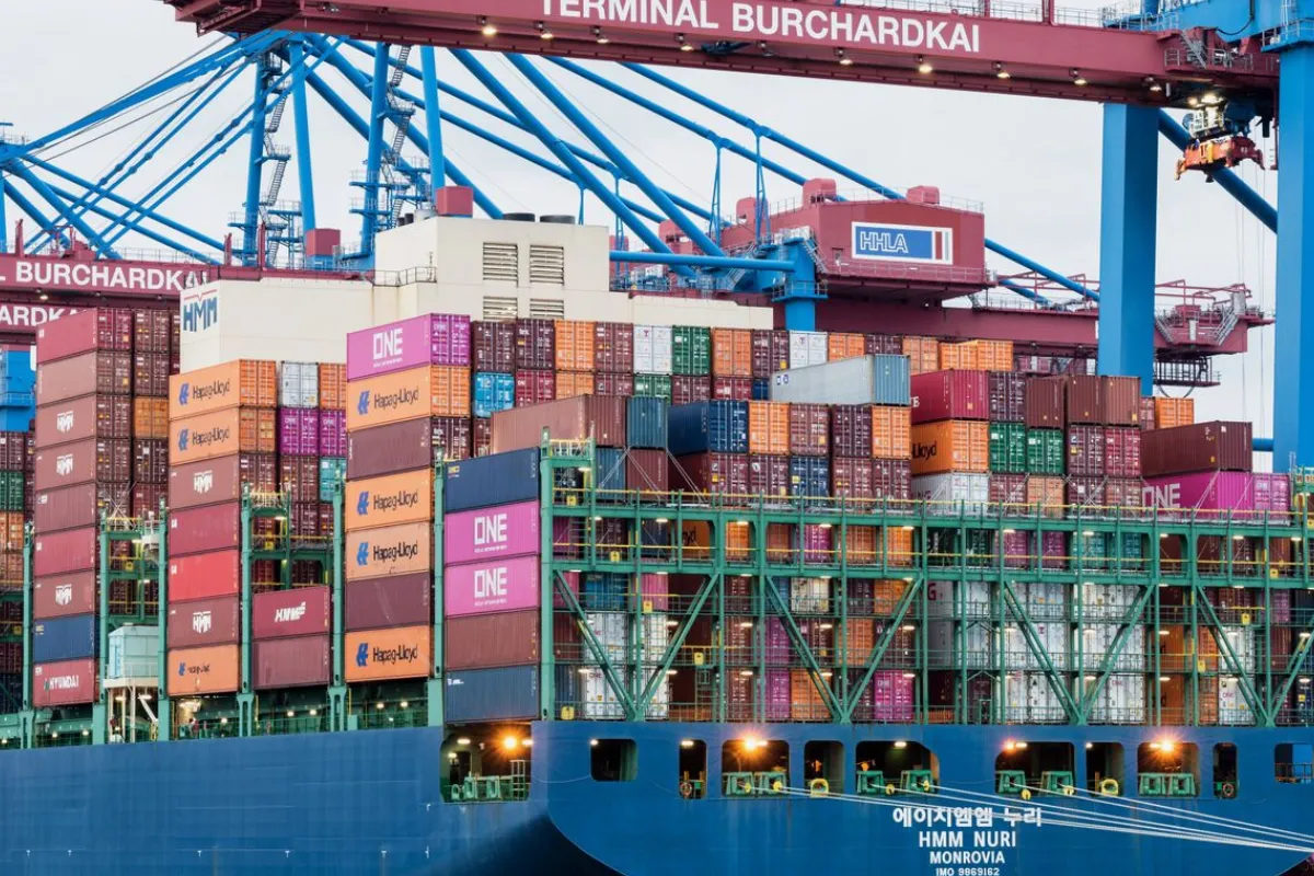 Navio porta-contêineres carregado atracado em terminal portuário com estruturas de movimentação de cargas.