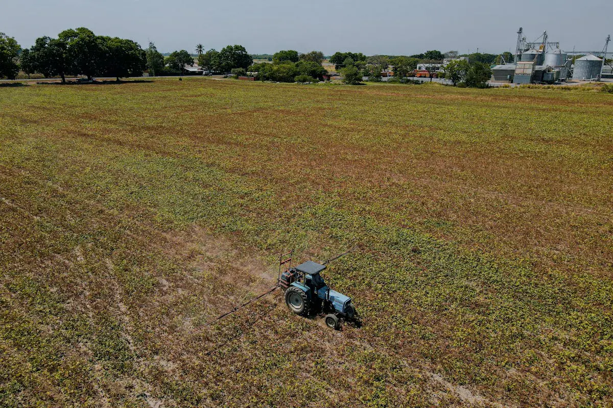 Vista aérea de um trator azul com barras pulverizadoras estendidas aplicando defensivos em um imenso campo de cultivo de tons esverdeados e marrons, tendo ao fundo silos metálicos industriais e árvores.