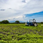 Vista em plano aberto de um trator azul e branco estacionado em um campo agrícola verdejante e irregular, sob um céu azul com nuvens brancas e montanhas distantes ao fundo.